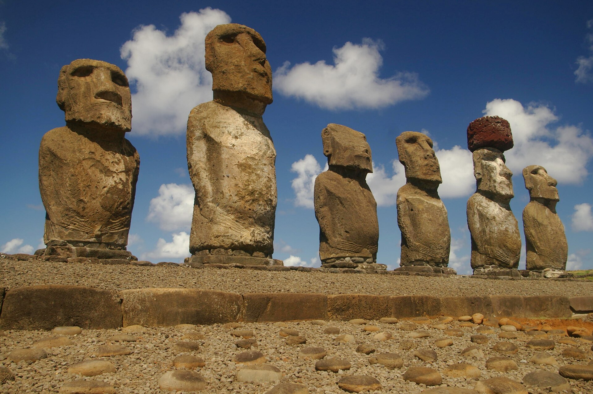 Seven moai standing on a long stone platform against a blue sky with clouds, one wearing a red stone topknot; hands carved low on the belly of several figures; a cobbled forecourt in front.