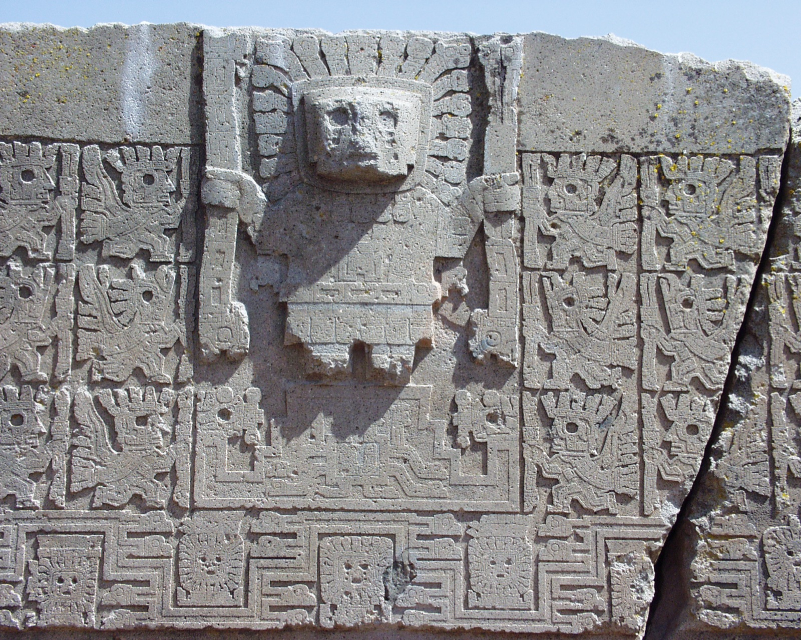 Detail of a carved stone lintel: a frontal figure with a rayed headdress holding a staff in each hand, flanked by rows of winged running figures in profile, above a meander border of small faces.