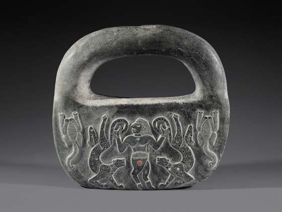 A dark chlorite object shaped like a handled bag, its face carved with a standing figure gripping a serpent on each side and further animals at the edges. A small photograph.