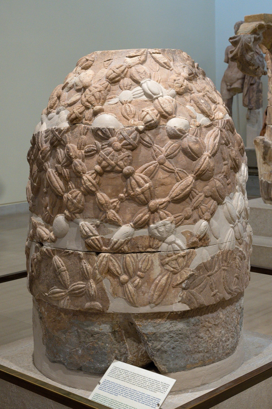 A conical marble stone covered in a carved net of knotted cords with bosses, standing in joined pieces on a museum floor beside a printed label.