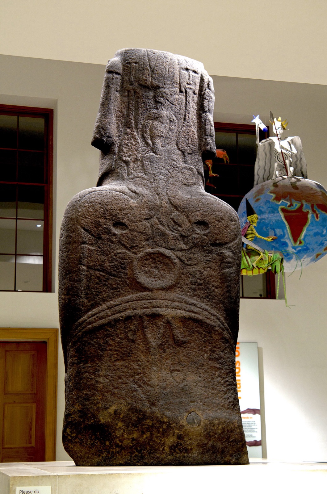 The back of a dark basalt statue in a museum gallery, carved in low relief: two facing bird-headed figures at the top, a disc within a ring-and-girdle motif at the waist, and paddle-like forms; a modern painted globe hangs in the background.