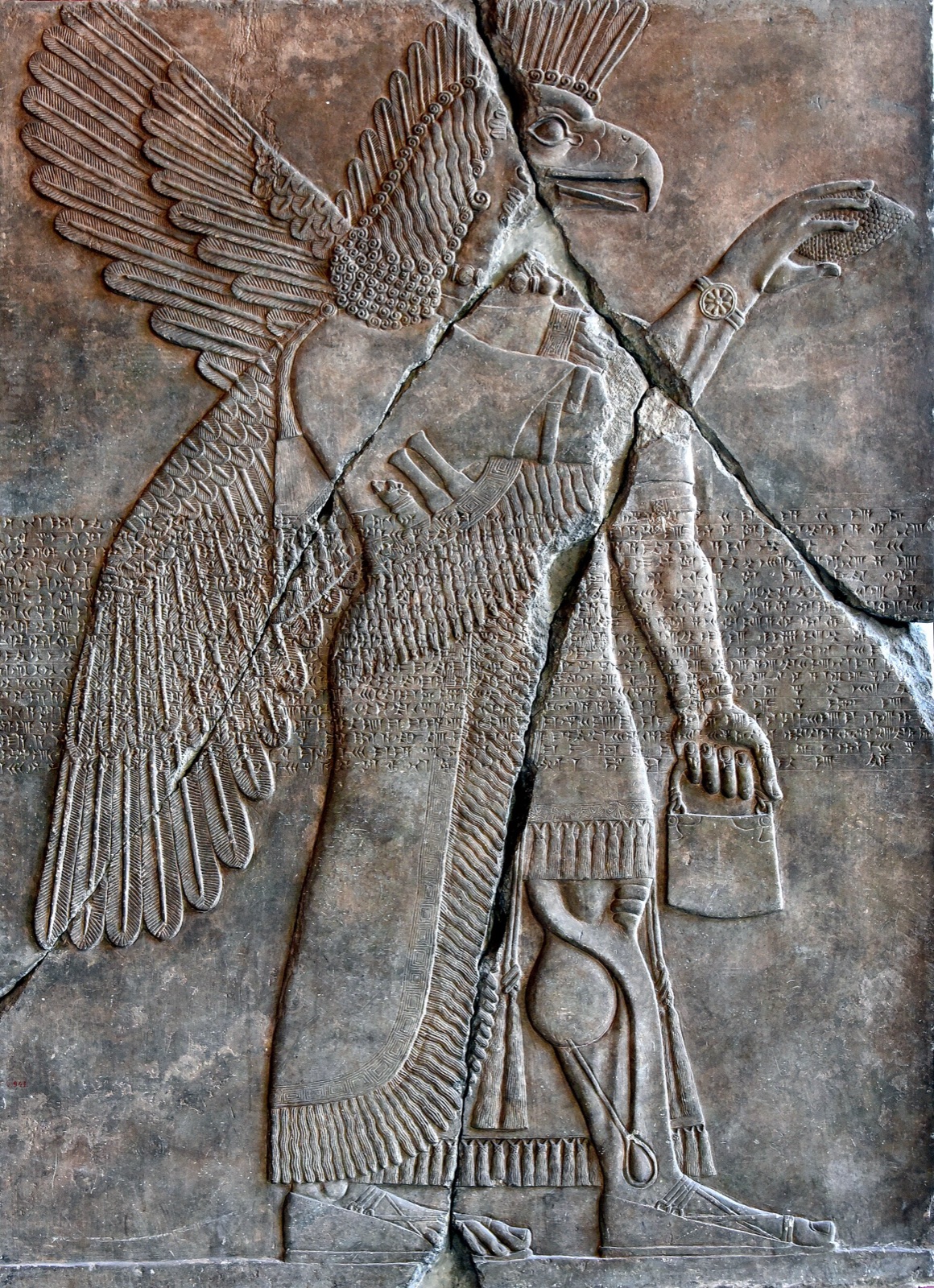 Alabaster wall relief: an eagle-headed, winged figure in profile striding right, a bucket hanging from the lowered hand and a cone raised in the other, with lines of cuneiform cut across the middle of the panel.