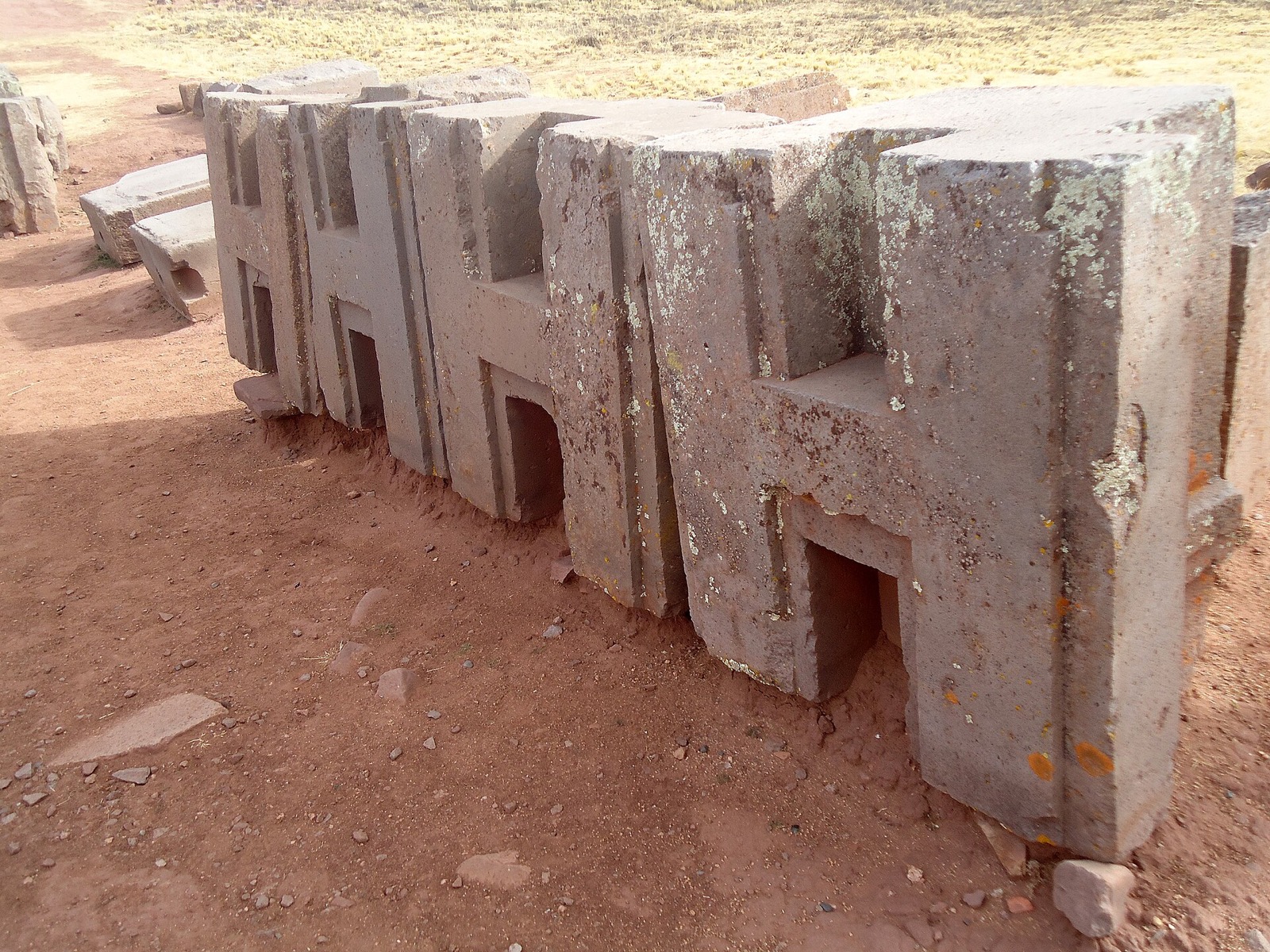 A row of grey andesite H-blocks at Puma Punku, each with identical stepped recesses and sharp inner corners.