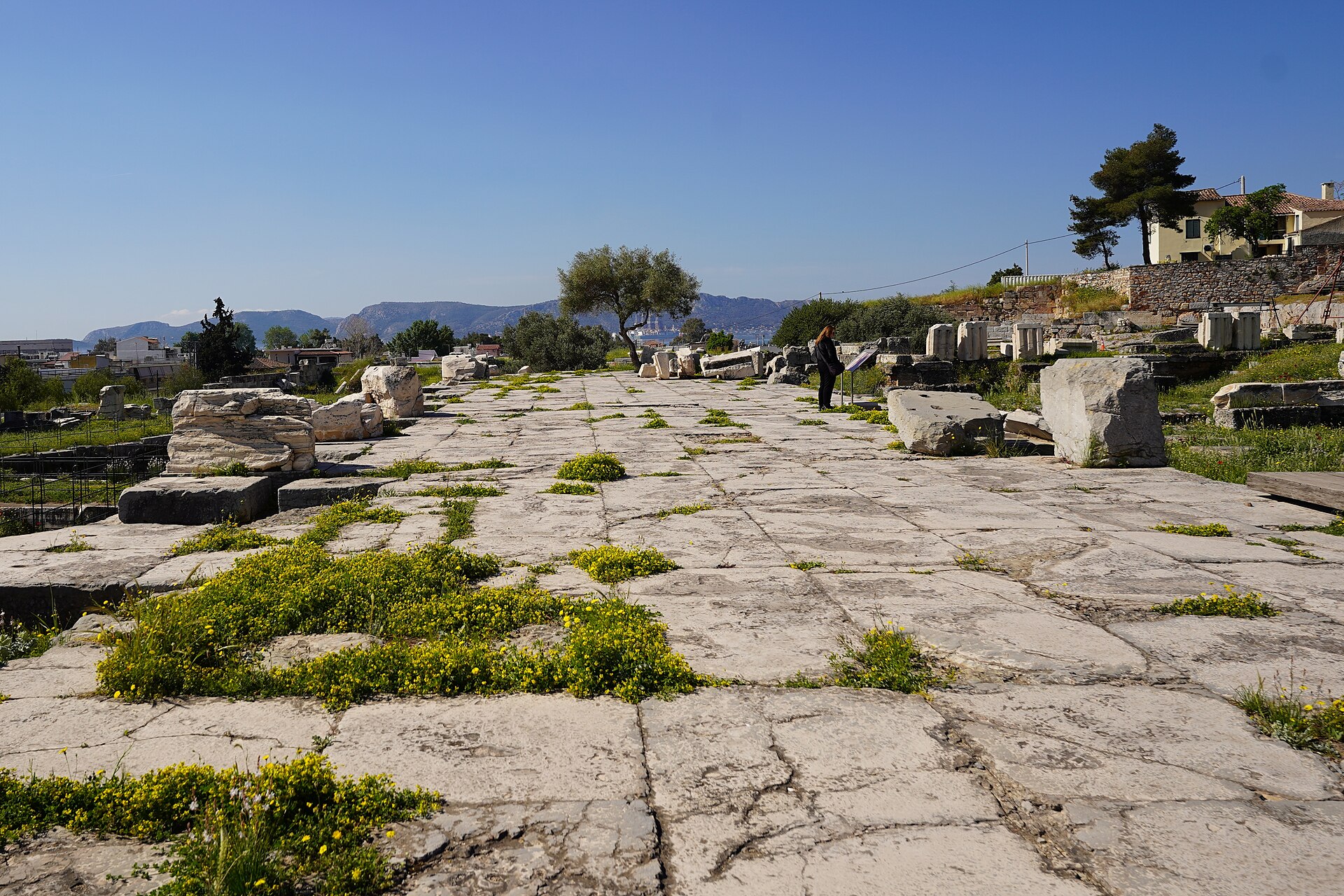 Broad expanse of weathered stone paving scattered with fallen marble blocks under a clear sky, a single visitor standing among them, with hills and the modern town of Elefsina beyond.