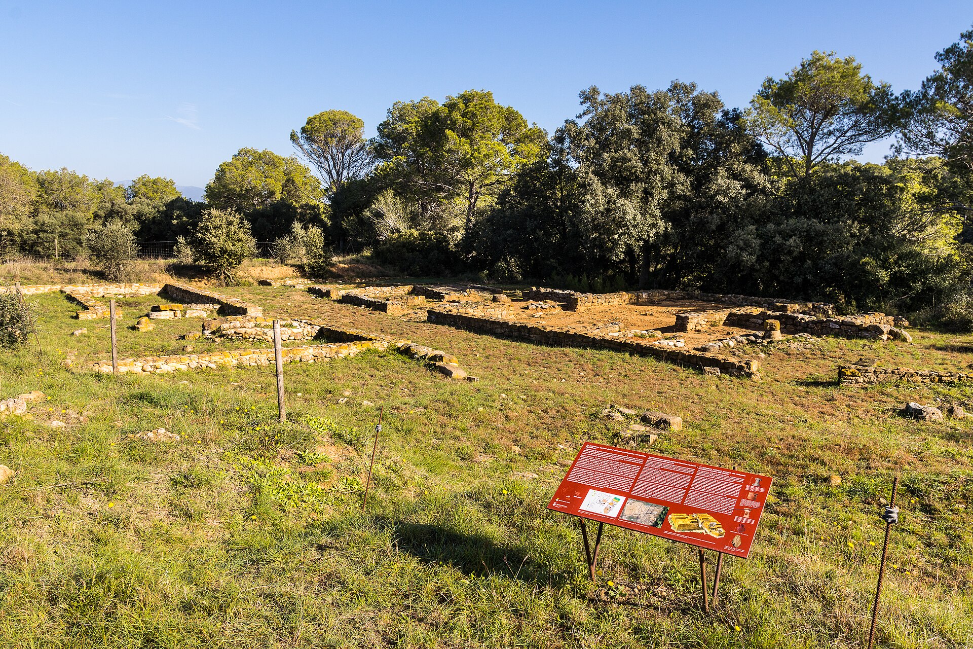 Low stone wall foundations of an ancient settlement in a grassy clearing ringed by trees, with a red interpretive panel in the foreground.