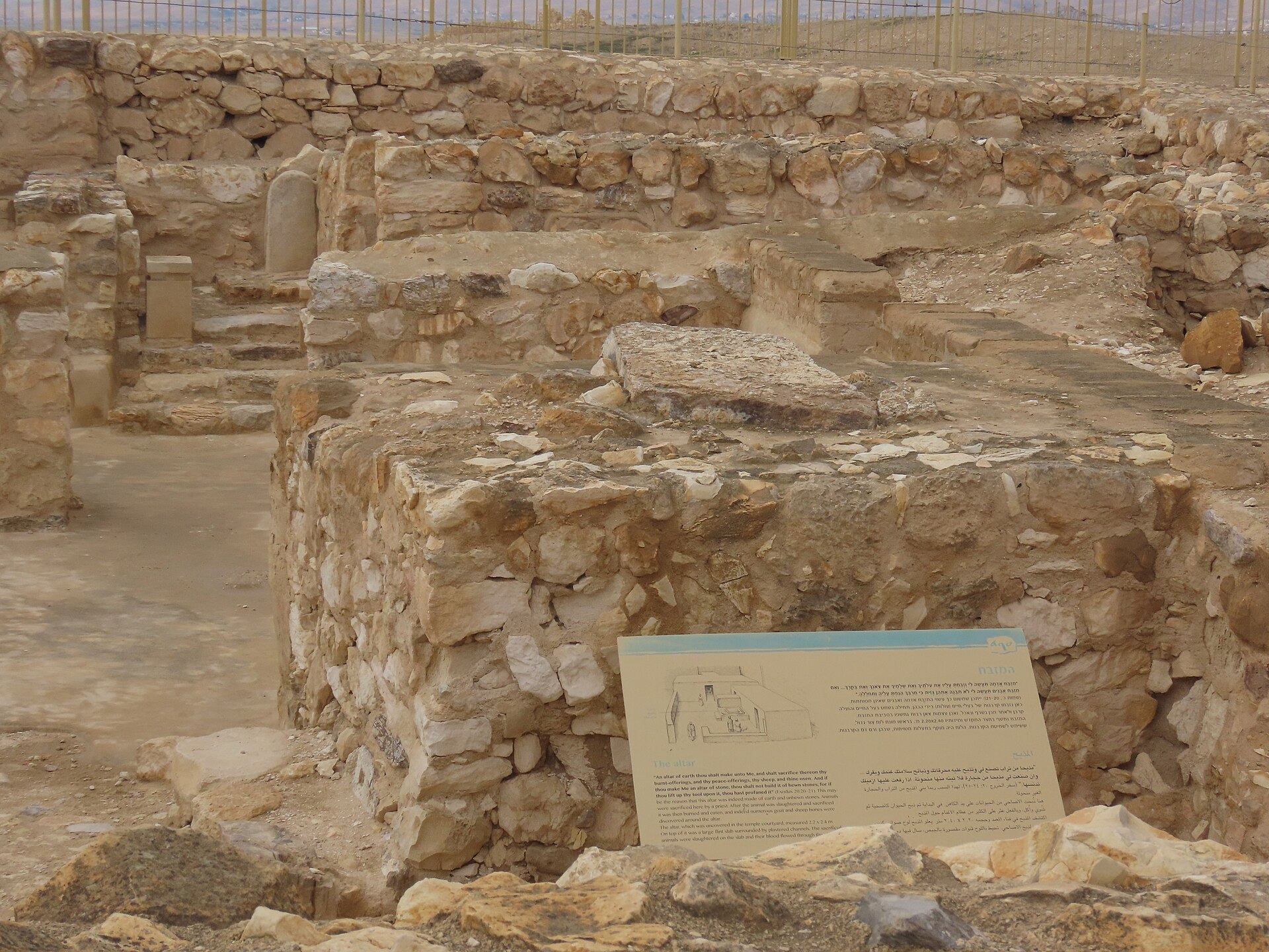 Stone ruins of a small temple at Tel Arad under an overcast sky: a large rubble-built altar block in the foreground and, behind it, steps rising to a niche holding a standing stone, with an interpretive sign in front.