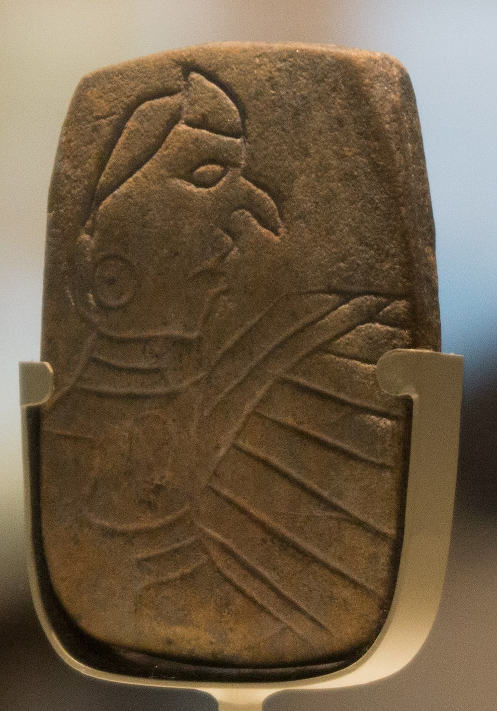 A small stone tablet on a museum mount, incised with a beaked human profile above a wing